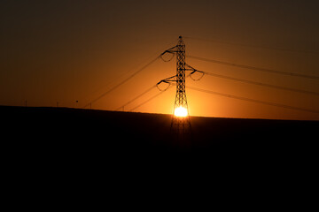 The sun hiding in the middle of an electricity tower with some mountains in the background against the light. The tower is in the middle of the picture.