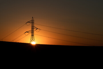 The sun hiding in the middle of an electricity tower with some mountains in the background against the light. The tower is on the left of the image.