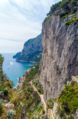 Fototapeta na wymiar A magnificent landscape from Giardini di Augusto in Capri, Italy. Mountains, rocks, little road and the boat passing by in the background, followed by the clouds and sky.