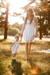 Fototapeta premium young beautiful mom is walking with little daughter in the sunny park . The concept of summer holiday outdoors. Mother's, baby's day. Spending time together. Sun light.