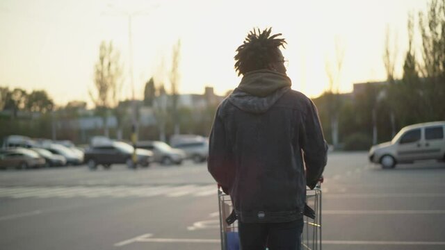 Back view of young African American man pushing shopping cart on parking at sunset. Camera follows guy in eyeglasses with dreadlocks strolling outdoors. Lifestyle.