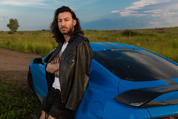 Stylish brutal man with long hair in a black leather jacket stands next to a bright blue sports car in the middle of the field against a clear sky on a warm day. High quality photo