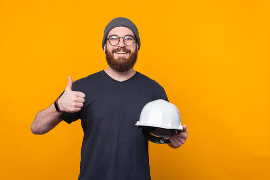 Photo of cheerful bearded man showing thumb up and holding helmet for construction.