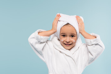 cheerful child in bathrobe and cosmetic cream on face adjusting towel on head isolated on blue