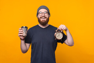 Portrait of joyful bearded hipster man holding alarm clock and coffee cup, its time to relax.