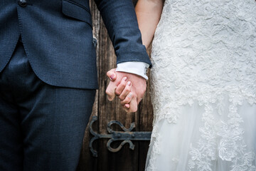 Close up of bride in white dress and groom in suit just married holding hands together following wedding ceremony stood in front of a wooden church door