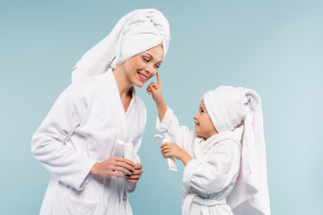 happy kid in bathrobe holding tube while applying cosmetic cream on face of mother isolated on blue