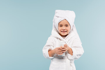 happy kid in bathrobe and towel on head smiling while holding tube with cosmetic product isolated...
