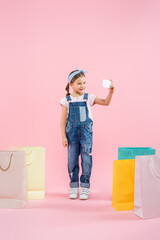 full length of cheerful kid taking selfie on smartphone near shopping bags on pink