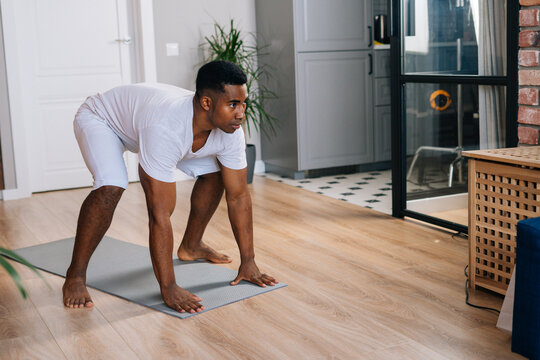 Strong African-American man practicing burpee exercise at home, doing push-ups and jumping on yoga mat at bright domestic room. Concept of sport training at home gym.