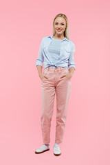 full length of happy and blonde woman smiling while standing with hands in pockets on pink