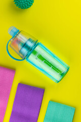 Colorful fitness gums on yellow background. Elastic expanders and tapes of different color. Colored rubber and textile bands, bottle of water and massage myofascial ball. Home fitness equipment.