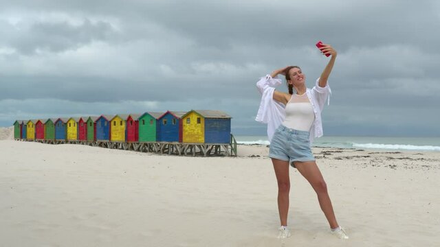 A girl make selfie on the white sands of South Africa near the colorful wood Muizenberg Beach Houses.