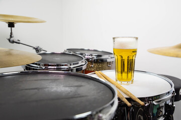 Glass of light beer on professional drum set closeup. Drumsticks, drums and cymbals, at live music rock concert, in the club stage, bar, or in recording studio. White background.