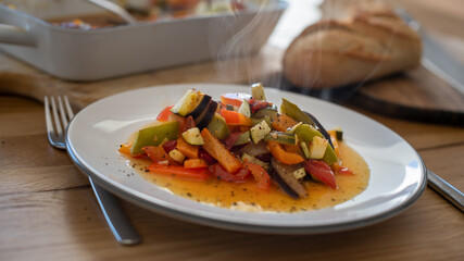Heißes dampfendes Rattatuille auf weißem Teller mit Baguette - rustikal auf einem Holztisch angerichtet