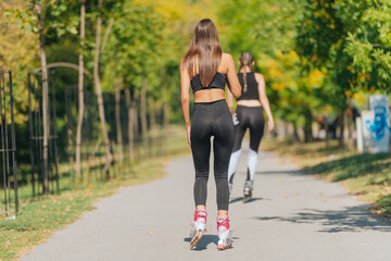 Rearview of two young fit women on roller skates riding outdoors on urban street in the park. - obrazy, fototapety, plakaty