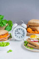 Alarm clock, healthy sandwiches with whole grains bread, microgreens, fresh vegetables Ketogenic diet, intermittent fasting, weight loss. Breakfast and dieting time concept. White table, copy space.