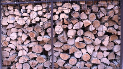 textured natural background of a fragment of a woodpile from large logs stacked behind a mesh metal fence, a street firewood warehouse in a village as a country style backdrop full frame