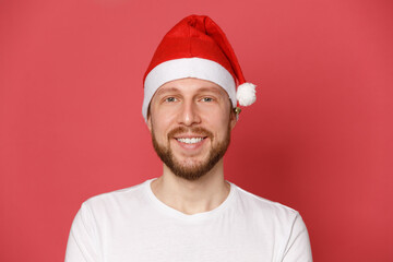 Portrait of a bearded guy in a white t-shirt in a santa claus hat over red pink background