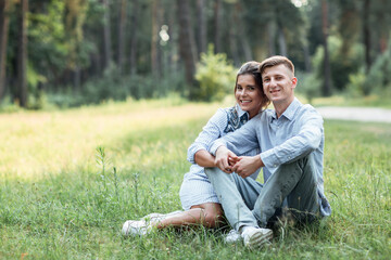 Naklejka na ściany i meble Outdoor shot of young happy couple in love sitting on grass on nature. Man and woman hugging, sunlight in summer park. Happy family in the evening sun light. The concept holiday