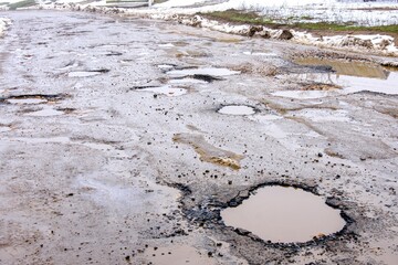 Road is paved with cracks in pits and puddles from the snow. Landscape of the winter road in cloudy gray weather. Concept of road repair, opacity for the driver of the car