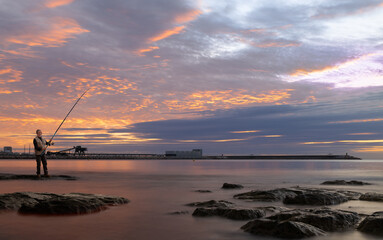 An angler stands on the rocks on the beach of Torrevieja in Spain in the early morning before sunrise. He has a fishing rod in his hand. In the background is the pier with the lighthouse.