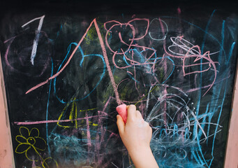Preschool girl child draws, writes with the right hand with pink chalk on a black wooden board,...