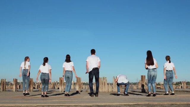 Young people in white T-shirts make a synchronized jump on the seashore, next to the pillars from the pier against the background of the blue sky. Shooting at 205 fps.