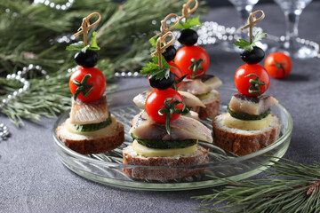 Canape with salted herring, cucumber, boiled potatoes, cherry tomato and black olives on rye croutons on gray background.