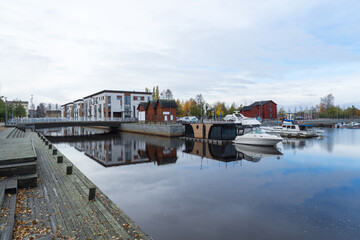 bridge to Kiikeli in Oulu with modern, clean and expensive apartments. Wooden pier with leaves, reflection in canal water. Houseboat and ships in harbor on beautiful autumn day