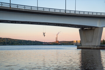 Kuokkalan silta bridge in Jyväskylä, Finland at lake Jyväsjärvi. Dangerous, adrenalin swing with person under bridge on a beautiful evening sunset with a smoking factory and clear finish lake water