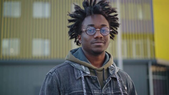 Portrait of African American man with dreadlocks in eyeglasses turning to camera smiling. Handsome young confident guy posing outdoors in urban city. Lifestyle concept.