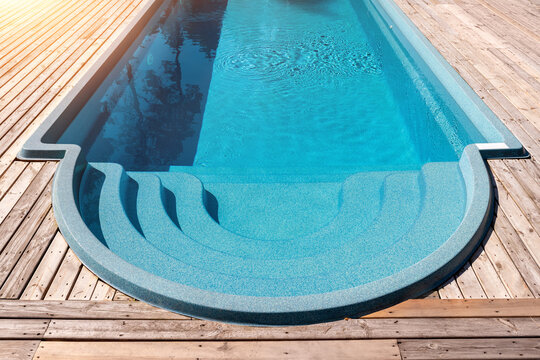 New modern fiberglass plastic swimming pool entrance step with clean fresh refreshing blue water on bright hot summer day at yard or resort hotel spa area. Wooden flooring deck of teak or larch board