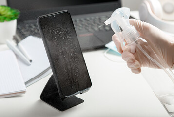 Woman in gloves uses antiseptic spray to clean smartphone on work space. Disinfection phone and laptop keyboard by alcohol disinfectant by woman in mask glowes on workplace, office desk
