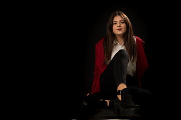 Mujer guapa y atractiva posa sentada sobre fondo oscuro vestida con camisa blanca chaqueta roja y pantalon negro con cara sonriente y alegre.