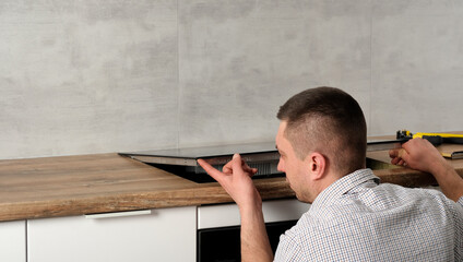 A young repairman installs a black induction hob in a modern white Scandinavian style kitchen with...