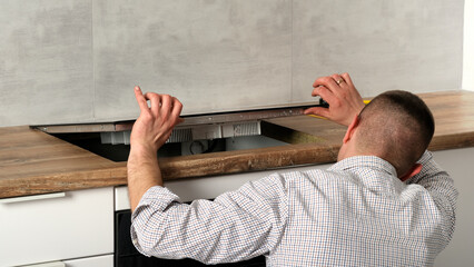 A young repairman installs a black induction hob in a modern white Scandinavian style kitchen with...