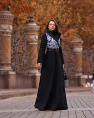 Elegant woman wearing black coat and walking city street on autumn or fall day against town park fence. Pretty girl with makeup and wavy brunette hair