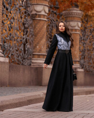Elegant woman wearing black coat and walking city street on autumn or fall day against town park fence. Pretty girl with makeup and wavy brunette hair