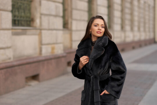 Elegant stylish woman with long brunette wavy hair wearing black trousers, pullover, shoes and fur coat walking city street on a sunny day