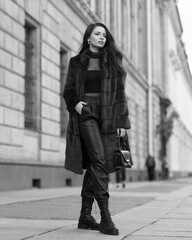 Elegant stylish woman with long brunette wavy hair wearing black trousers, pullover, shoes and fur coat walking city street on a sunny day