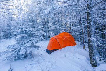 An orange tent stands among the snow-covered trees in the winter forest. Bright tourist tent in the winter taiga