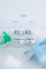 covid-19 vaccine against the pandemic, ampoules with Vaccine 1 and Vaccine 2 labels side by side next to 2021 yearly calendar and mask gloves and sanitizer