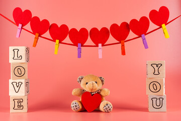 A teddy bear with a red heart. Wooden cubes with the inscription love you. A greeting card on a pink background. garland of red hearts on tape. Valentine's day background.