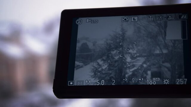 Watching the heavy blizzard in UK through a camera display, snow-covered houses and trees on the street. Exposition details and settings are displayed on the screen. Slow motion close up 4k video