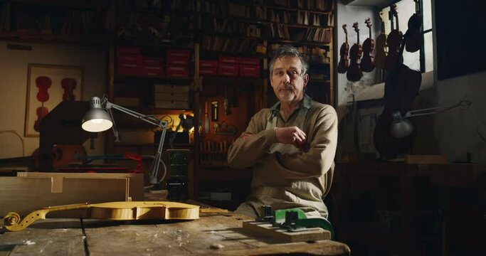Cinematic shot of professional master artisan luthier is smiling satisfied with his job in camera while working on creation of handmade fine quality wood violin in creative workshop.