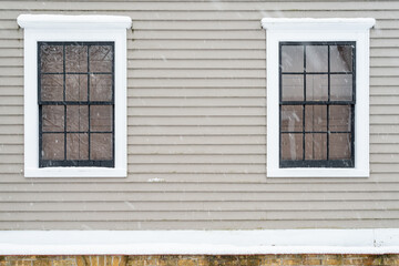 Fototapeta na wymiar Two vintage identical double hung windows with reflecting glass on a beige colour exterior wall. The windows are dark green with white trim. There's a rock foundation below the wood clapboard wall.