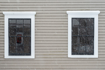 Two vintage identical double hung windows with reflecting glass on a beige colour exterior wall. The windows are dark green with white trim. There's a rock foundation below the wood clapboard wall.