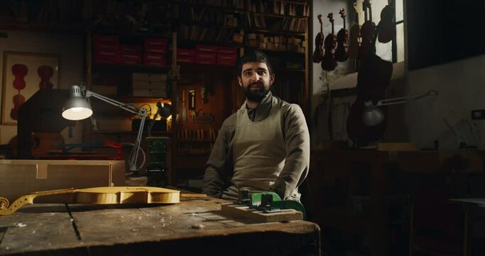 Cinematic shot of young experienced master artisan luthier is smiling satisfied with his job in camera while working on creation of handmade fine quality wood violin in creative workshop.