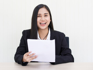 Fototapeta premium portrait of young beautiful and happy Asian woman wearing smart casual laughing with friendly and cheerful face holding paper sitting on a desk celebrate for her success